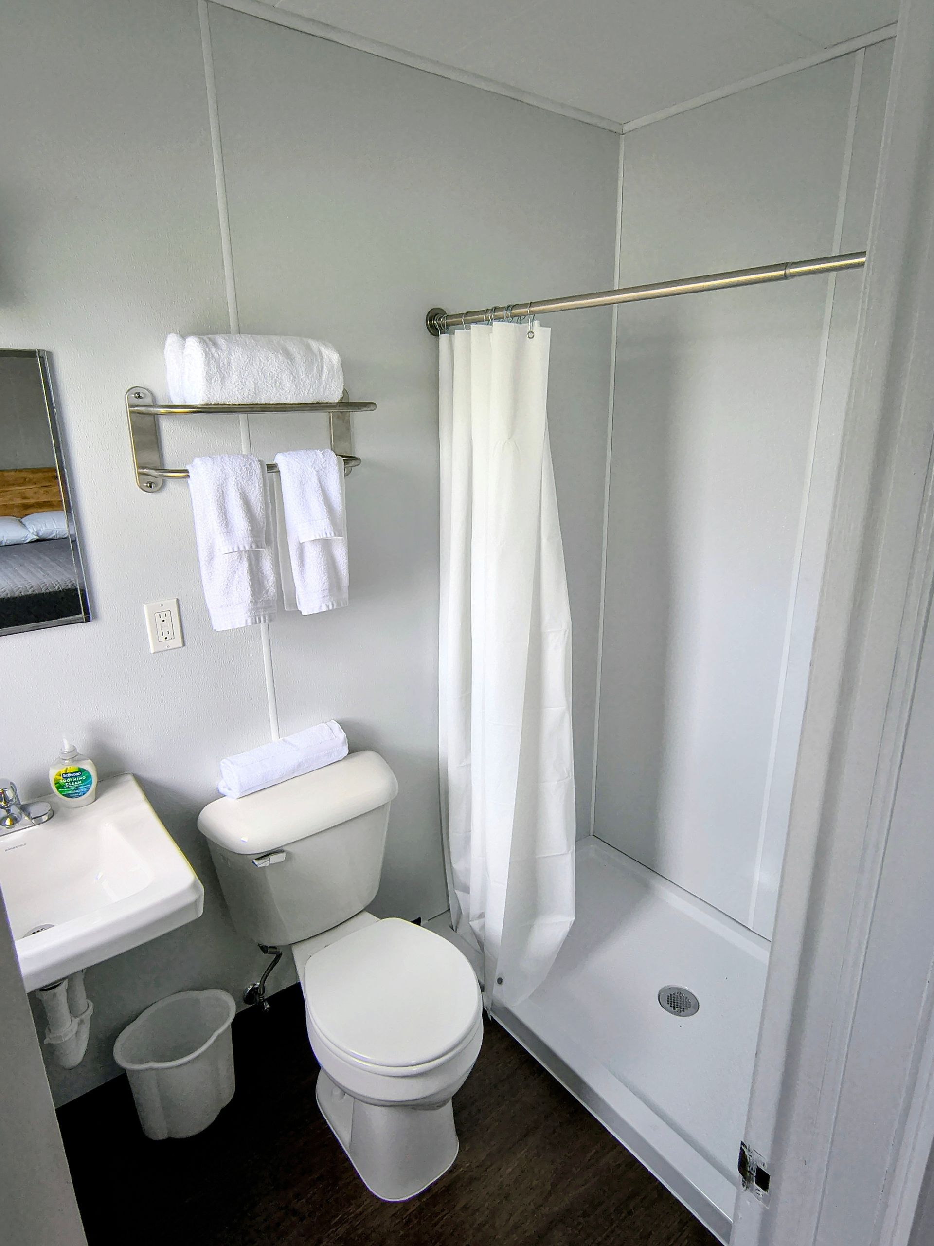 A small motel bathroom with a toilet, sink, and shower. White walls, dark floor, and white towels on a rack.