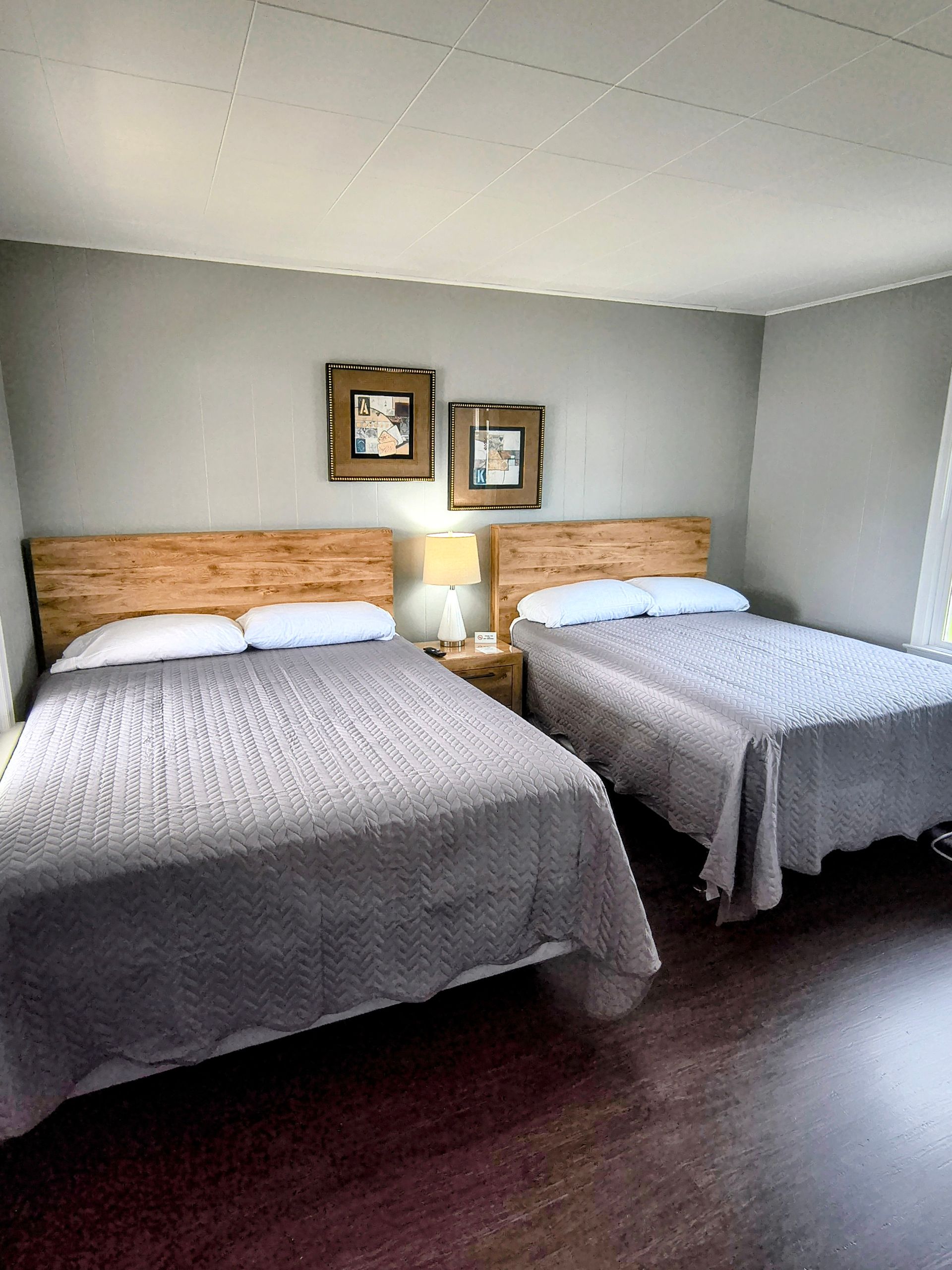 Two beds in a bedroom with wooden headboards, gray bedding, and framed art on a gray wall.