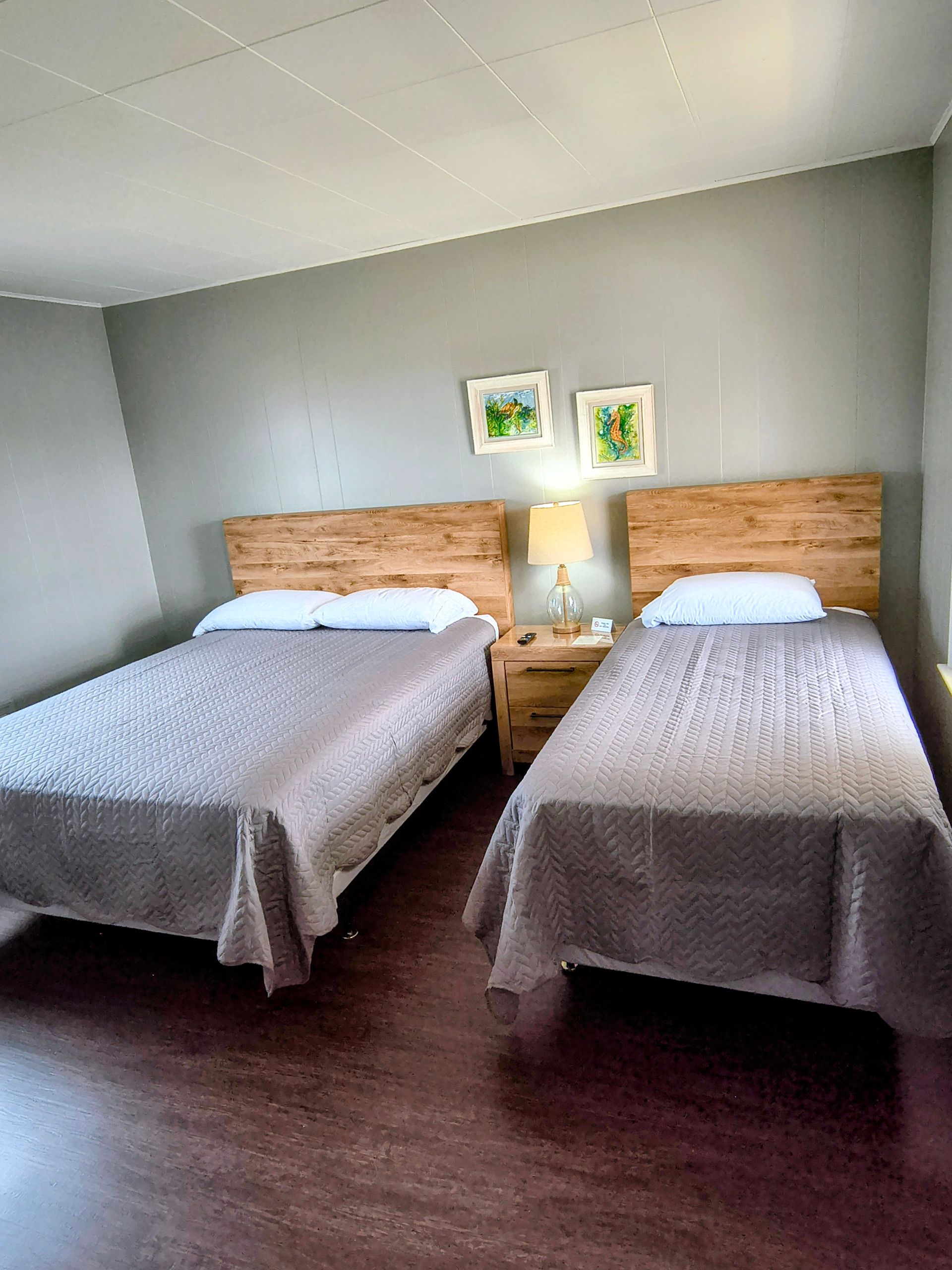A motel room with two beds. The beds have wooden headboards and gray bedspreads. Artwork hangs above a nightstand.
