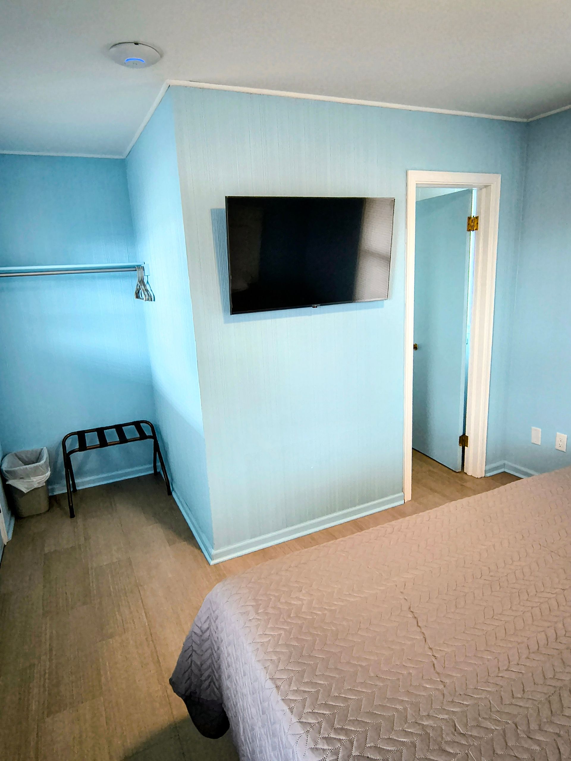 Light blue motel room with mounted TV, bed, closet, and a doorway.