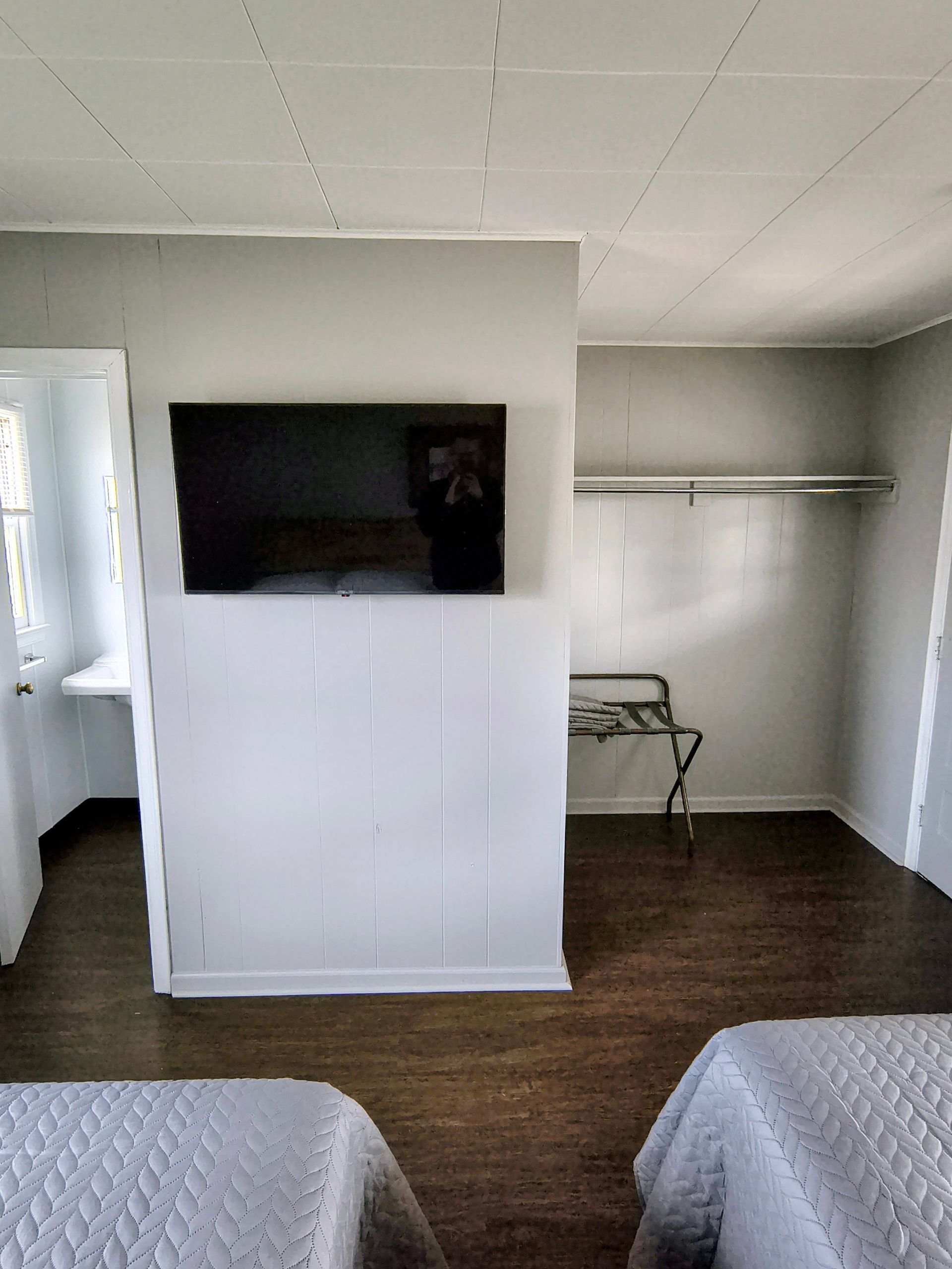 Bedroom with two beds, a wall-mounted TV, and a closet. Walls are light gray with dark wooden floors.