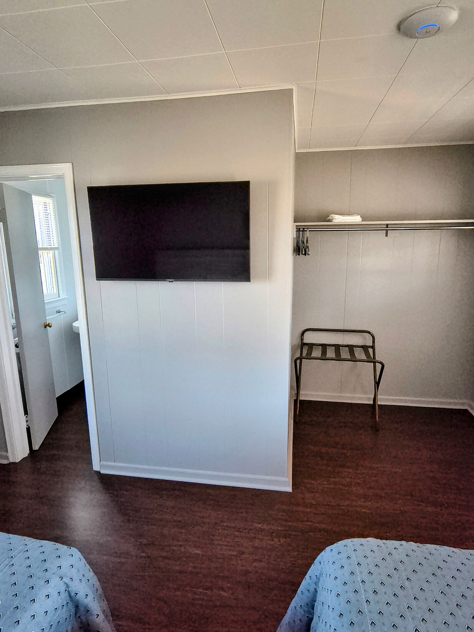 A motel room interior features a mounted TV, closet with hangers, and dark wood-like flooring.