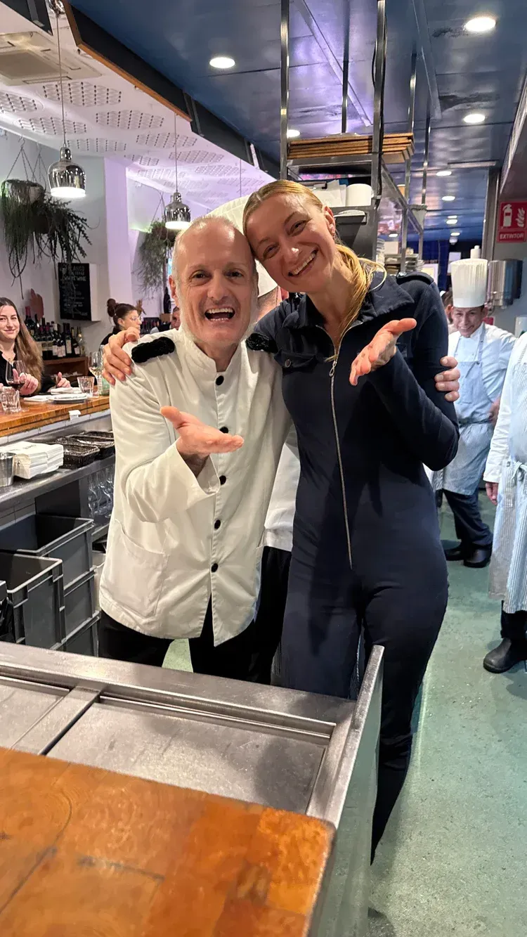 Two smiling people posing in a brightly lit restaurant, leaning together by a counter.
