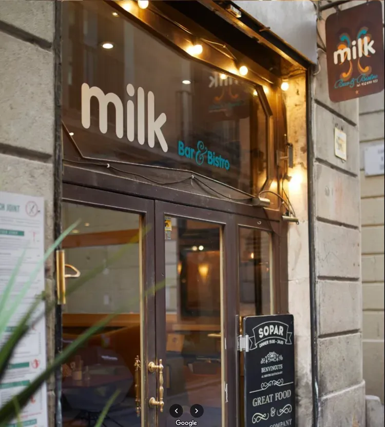 Small storefront with “milk” sign and glass entrance on a narrow street, warm lights glowing inside