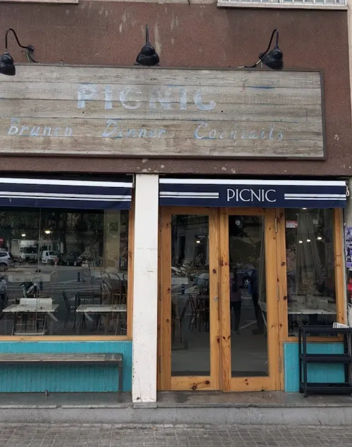 Storefront with faded wooden sign reading “PICNIC,” glass doors, and blue awning on a street.