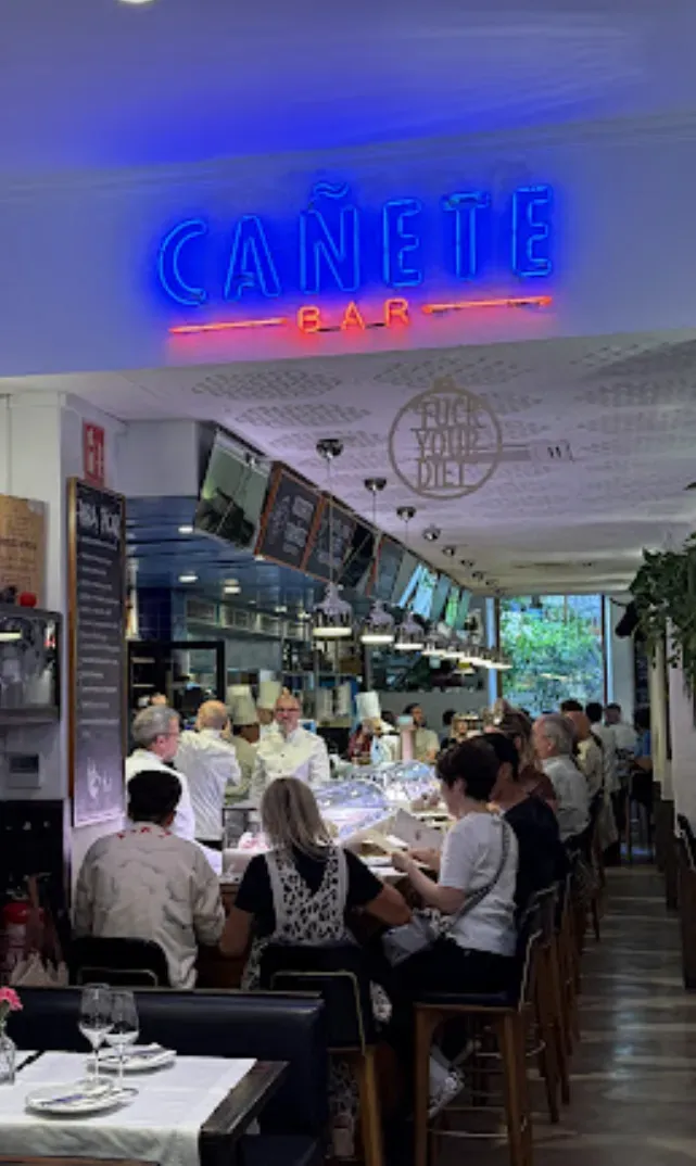Busy restaurant interior with diners at tables beneath a blue neon sign.