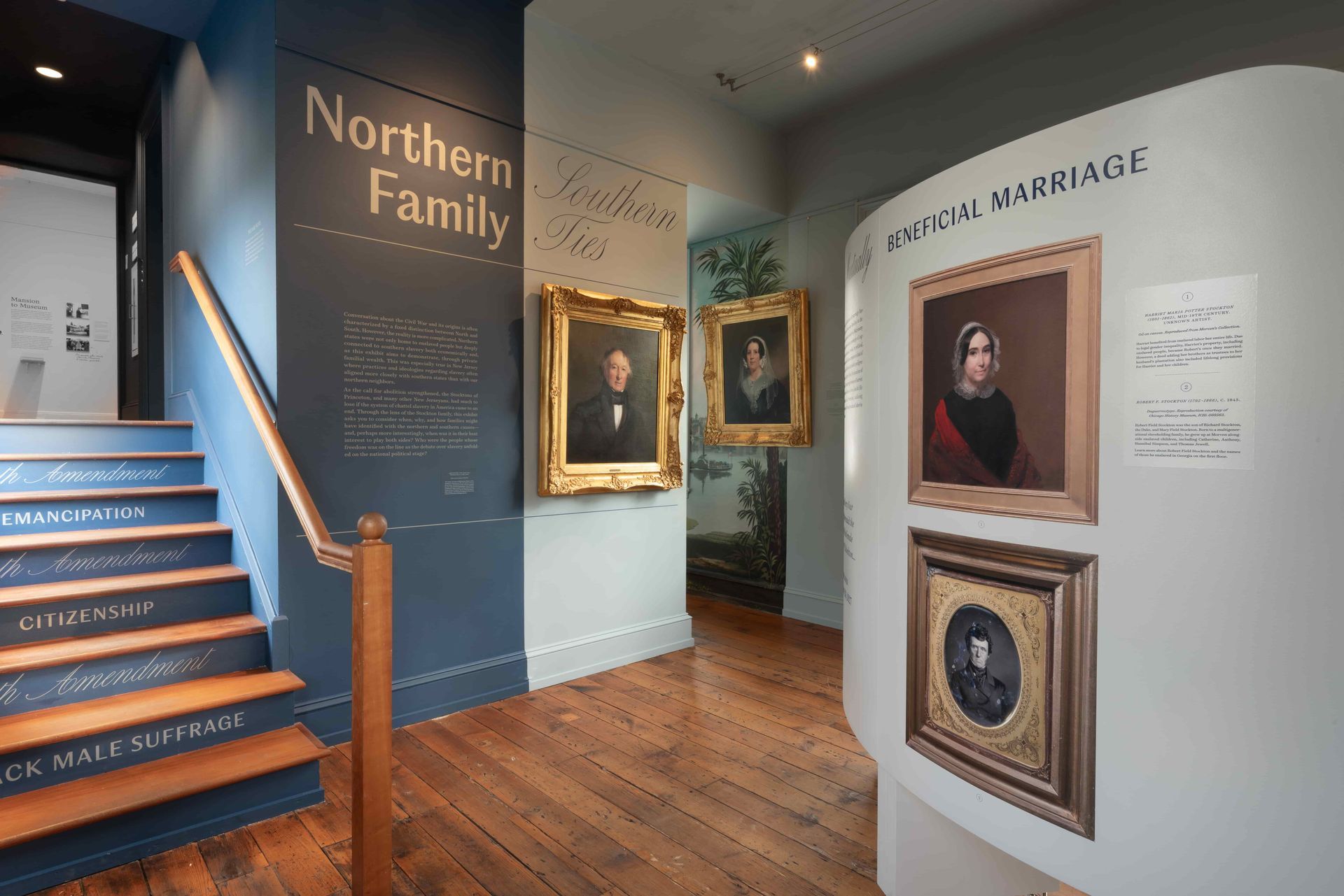 Museum interior, portraits in gilded frames on display, stairs, text panels, wooden floor, blue and white walls.