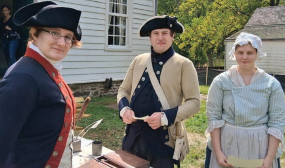 Two men and a woman dressed in everyday colonial garments, standing around a table outdoors.