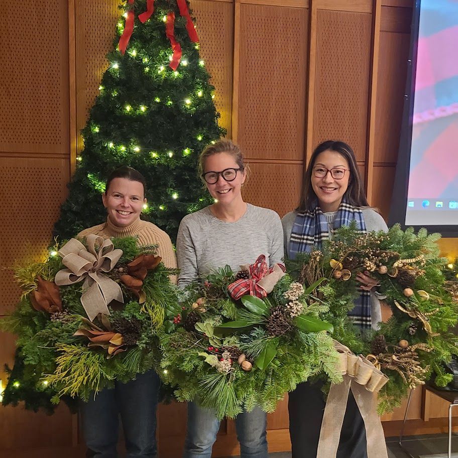 Three people holding holiday wreaths in front of a lit Christmas tree
