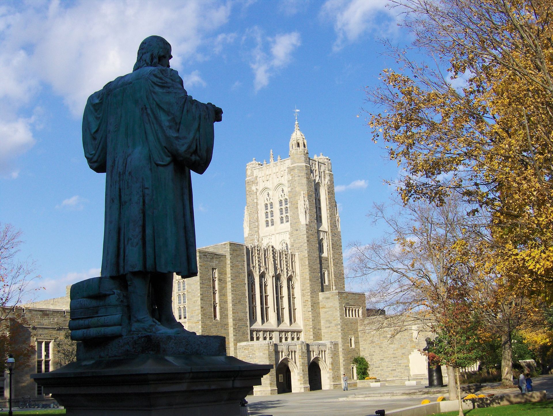 Statue in foreground facing a tall stone cathedral under a blue sky, with autumn trees nearby
