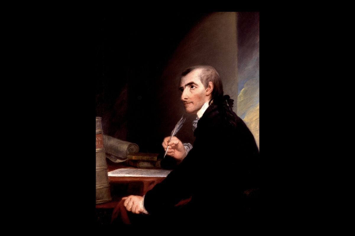 Portrait of a seated man in a dark suit, holding a paper beside a desk in a dimly lit room