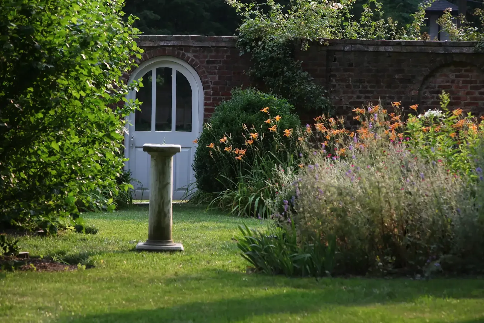 Garden with a a lush green lawn, flowering shrubs, and a white arched door in a brick wall.