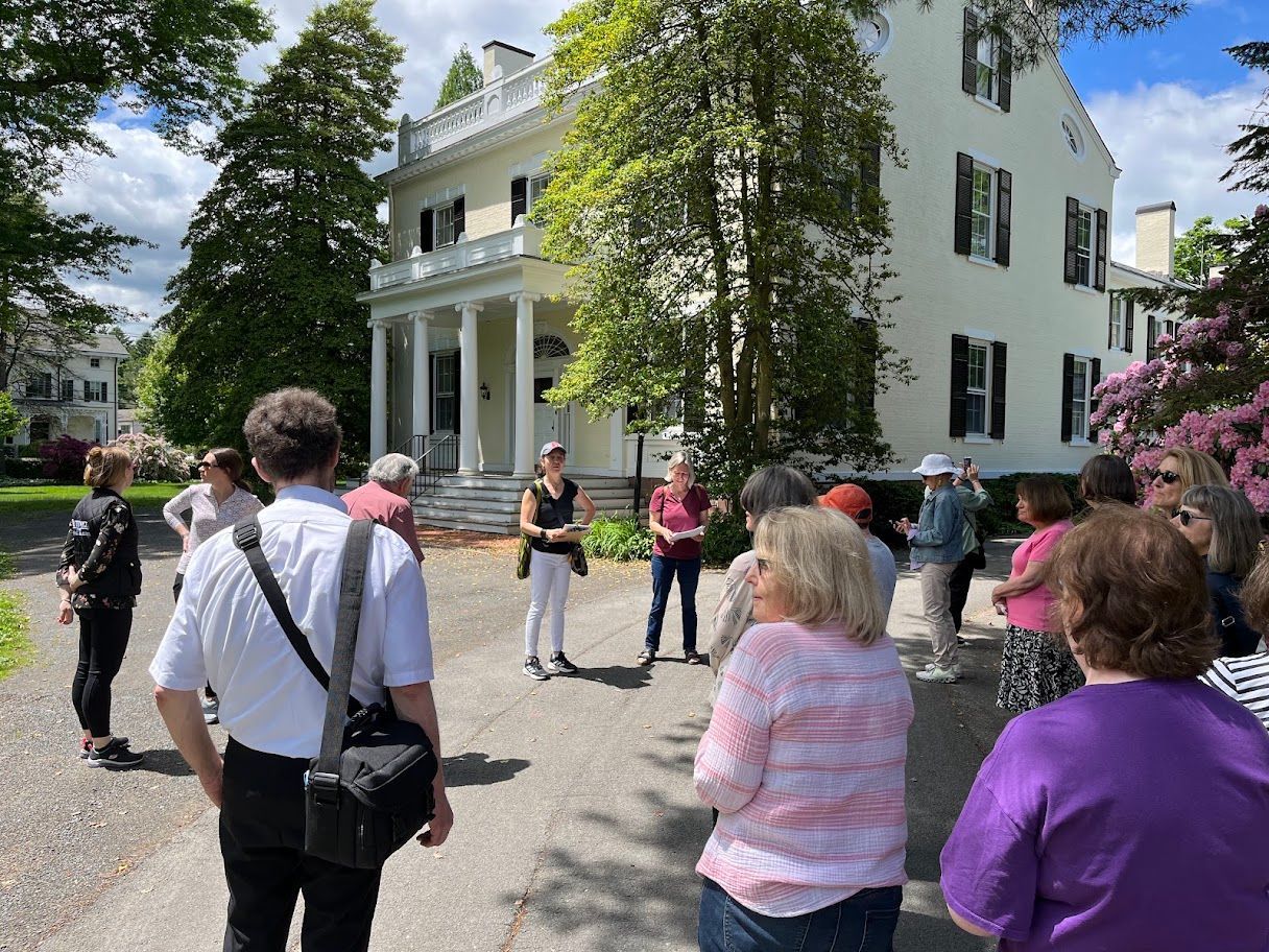 People gathered outside a white house with a porch on a sunny day.