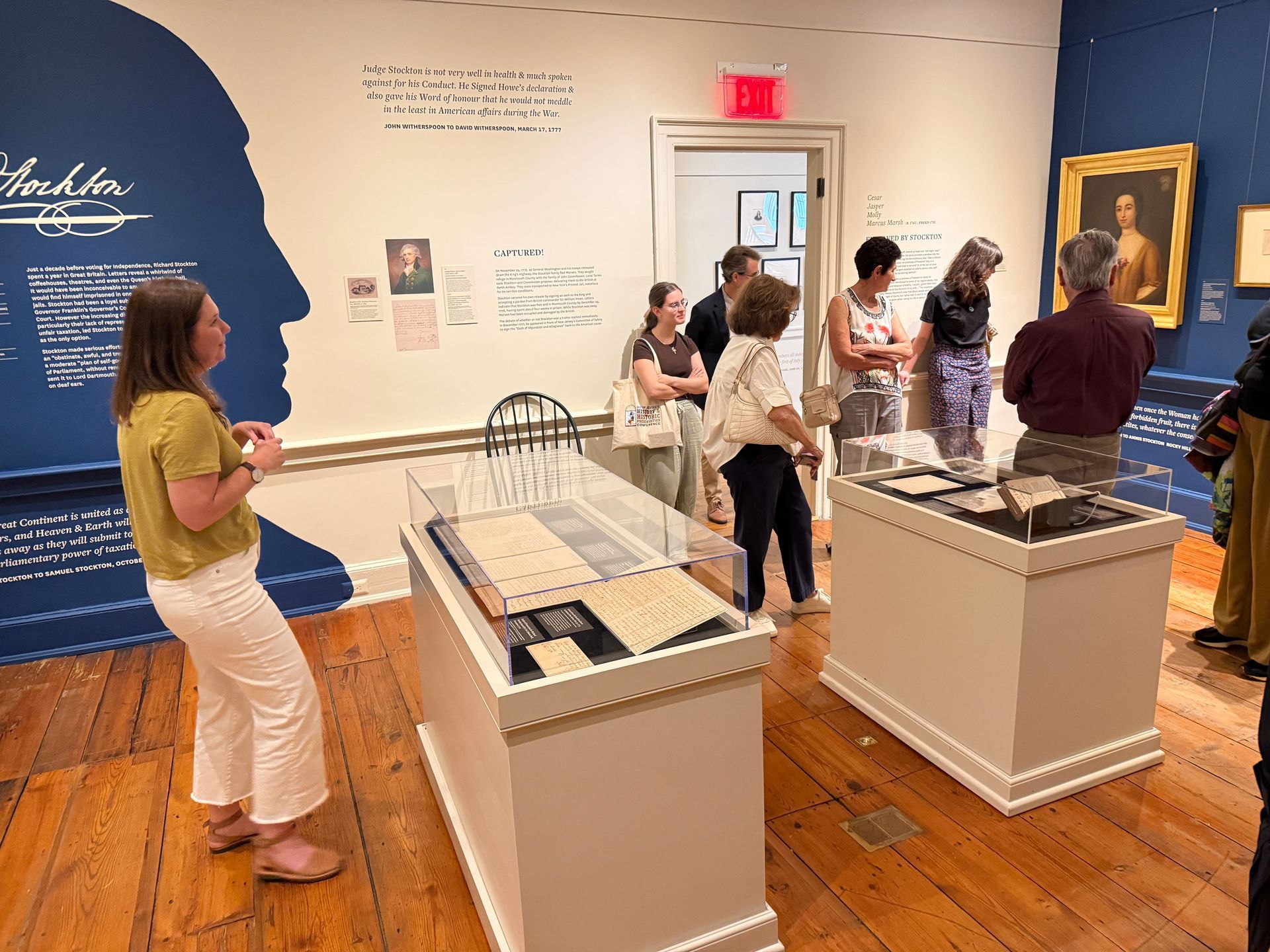 Museum exhibit room with visitors viewing display cases and wall text beneath blue and white gallery walls