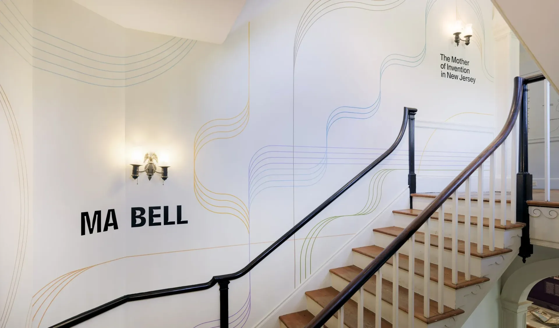 Staircase with "MA BELL" and map-like art on the wall, black handrail. Wooden stairs, two sconces.