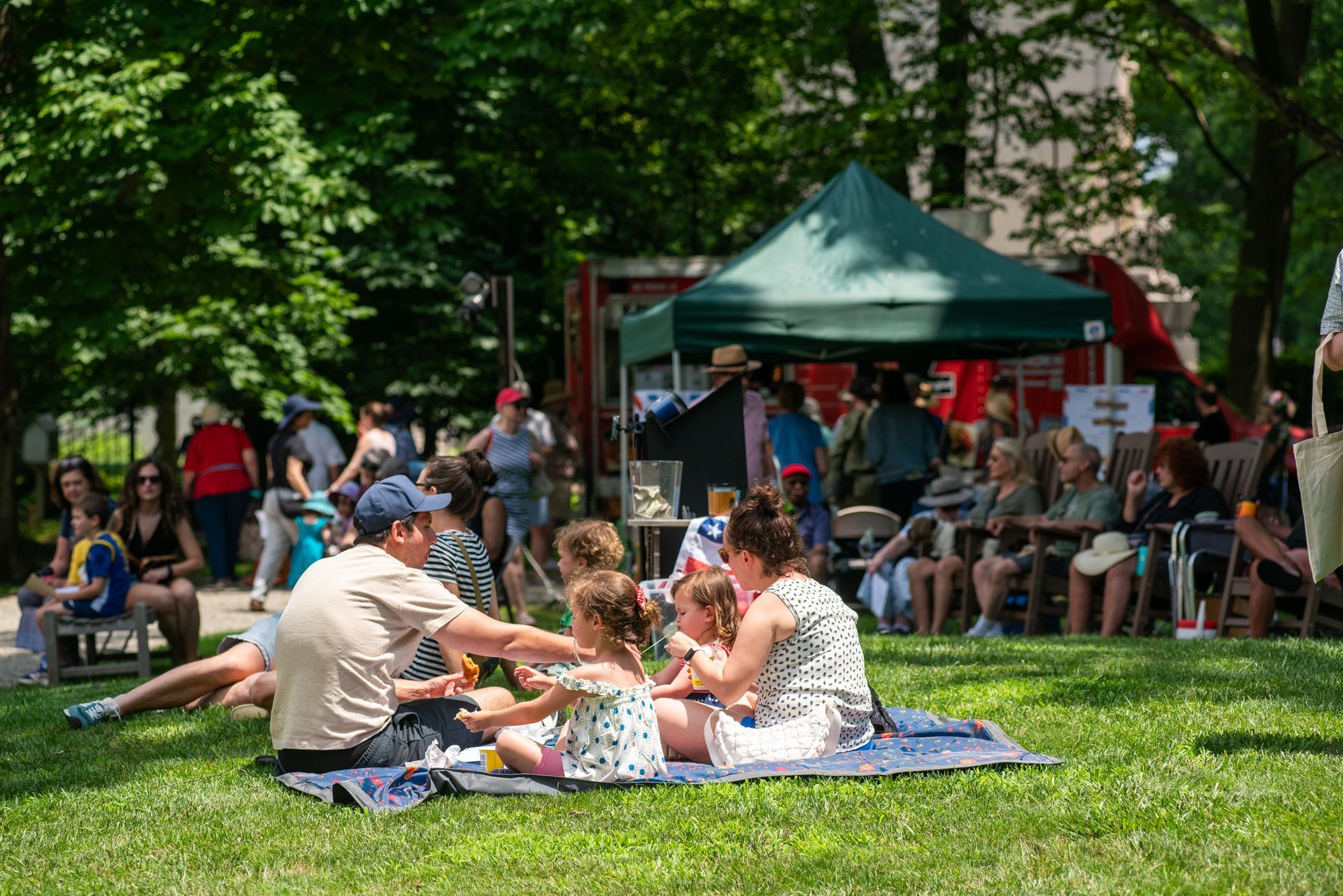 People sitting on blankets at a sunny park gathering with a green tent and crowd in the background