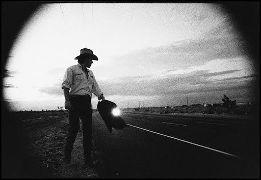 Man in cowboy hat holds boot on highway at dusk. Black and white.