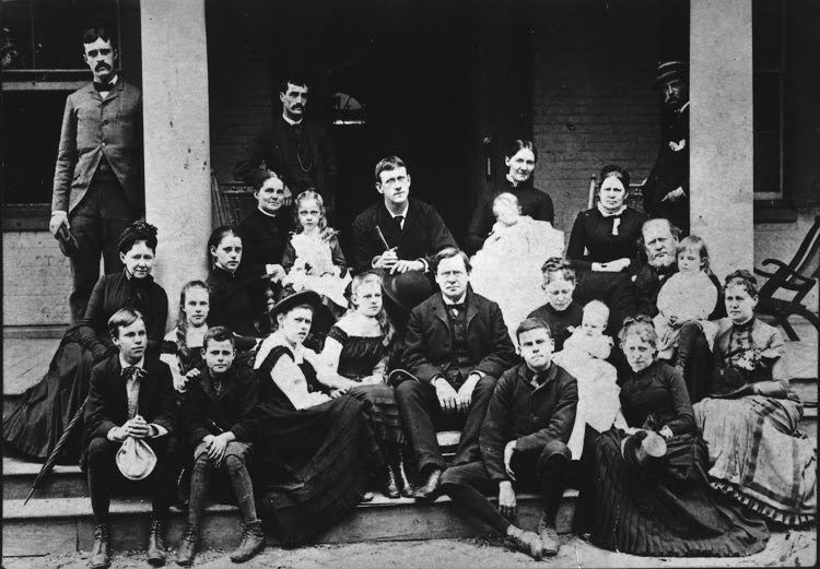 Family group posing on a porch. Men in suits, women in dark dresses, children. Black and white photo.