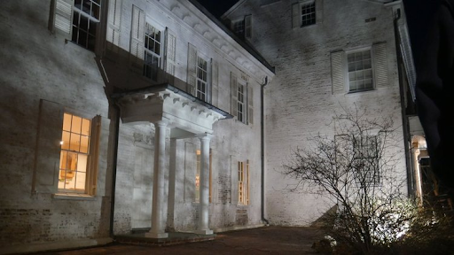 White colonial house at night, lit by spotlights, with lighted windows and bare tree.