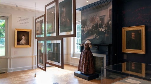 Museum exhibit, historical portraits and dress displayed. Interior with framed art, glass cases, and fireplace.