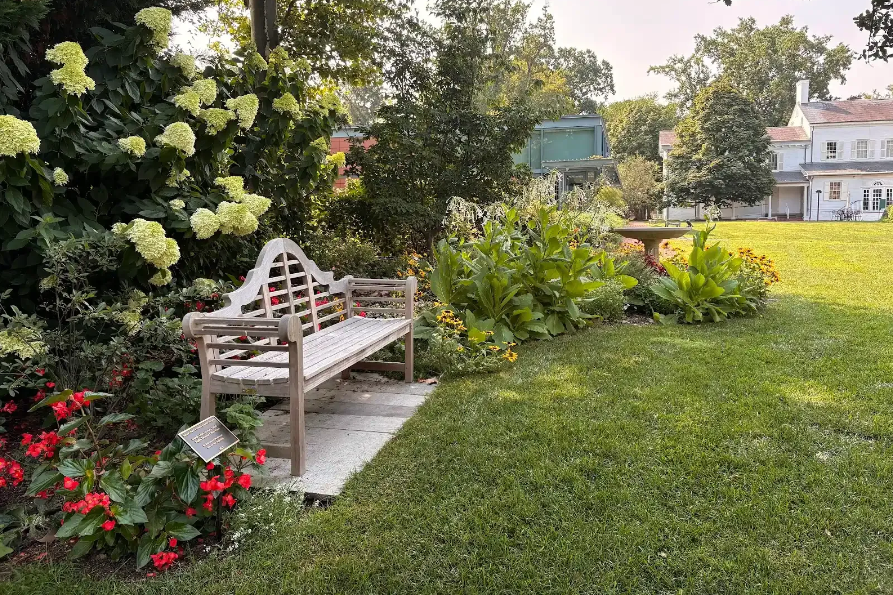 Wooden bench in a garden, with flowers and a white house in the background. Green grass and trees surround it.