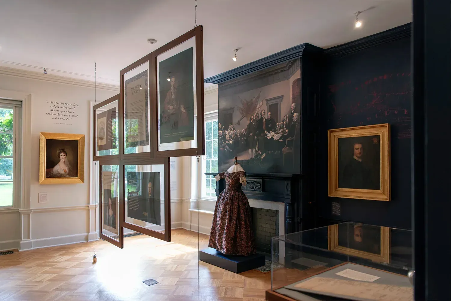 An art gallery with portraits and a dress on display. Light wood floors, dark blue walls, and framed art.