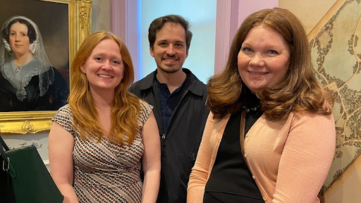Three people smiling in a room. A painting hangs on the left; a map is on the right.