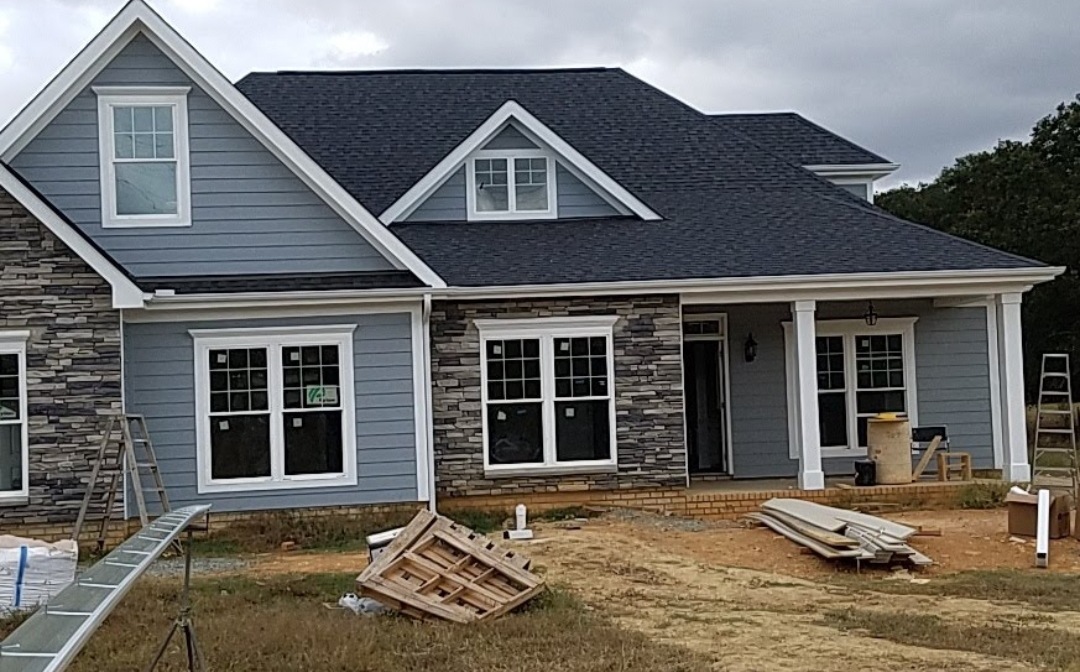 A house under construction with blue siding, stone accents, and a dark roof; tools and materials scattered around.