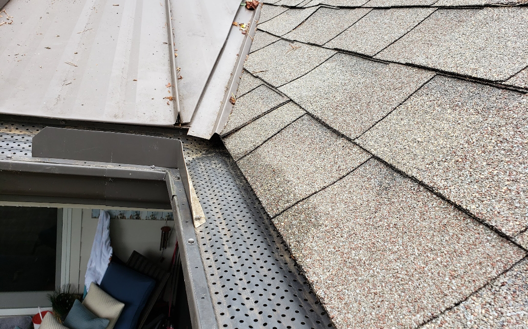 Close-up view of a house's roof and gutter, showing a gutter guard and weathered shingles.