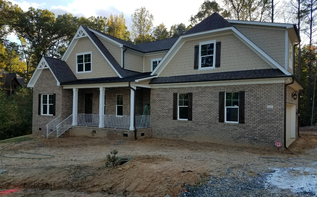 Two-story house under construction; tan siding and brick exterior, dark shutters, in a clearing, daytime.
