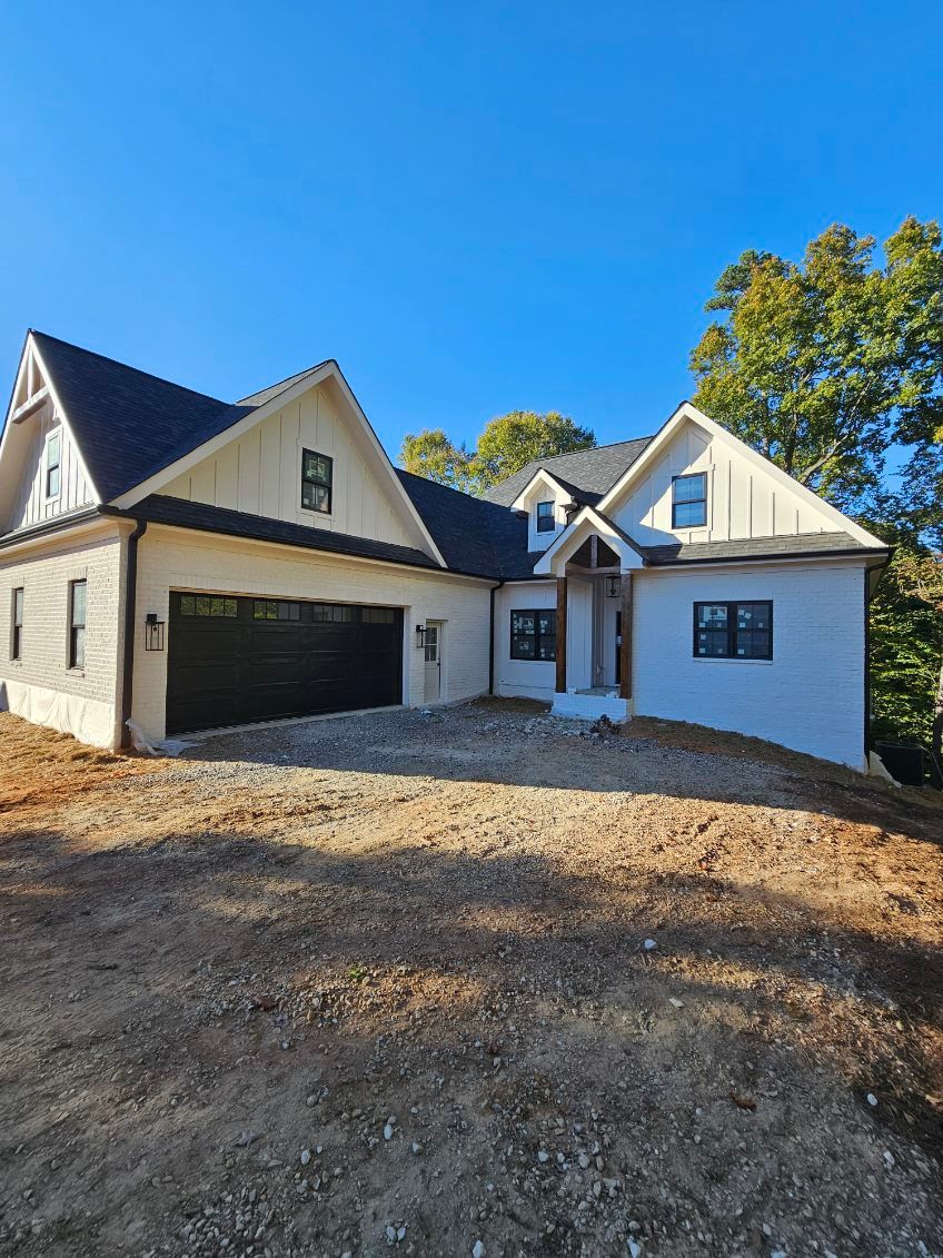 Newly constructed white house with black roof and garage door, on a dirt lot.