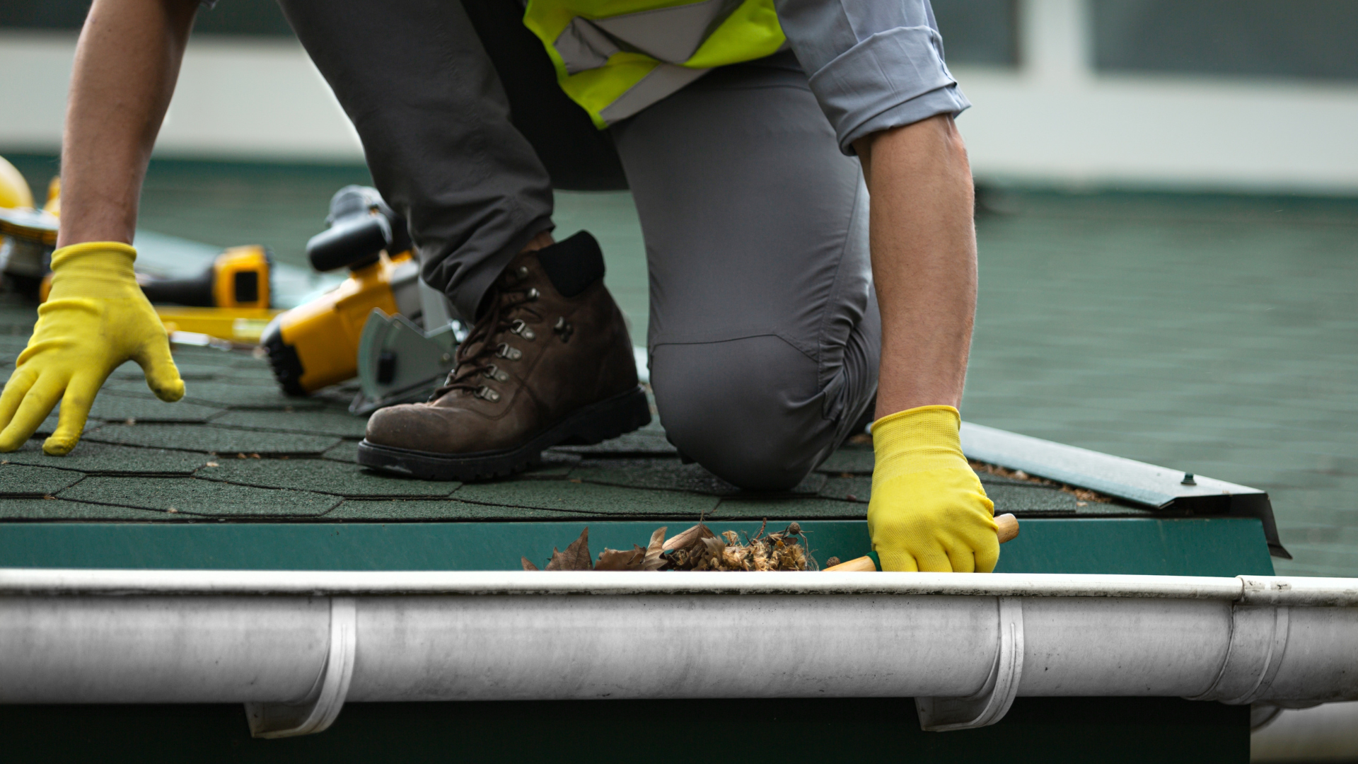 Person in safety gear cleaning a gutter on a roof, removing debris with yellow gloved hands.