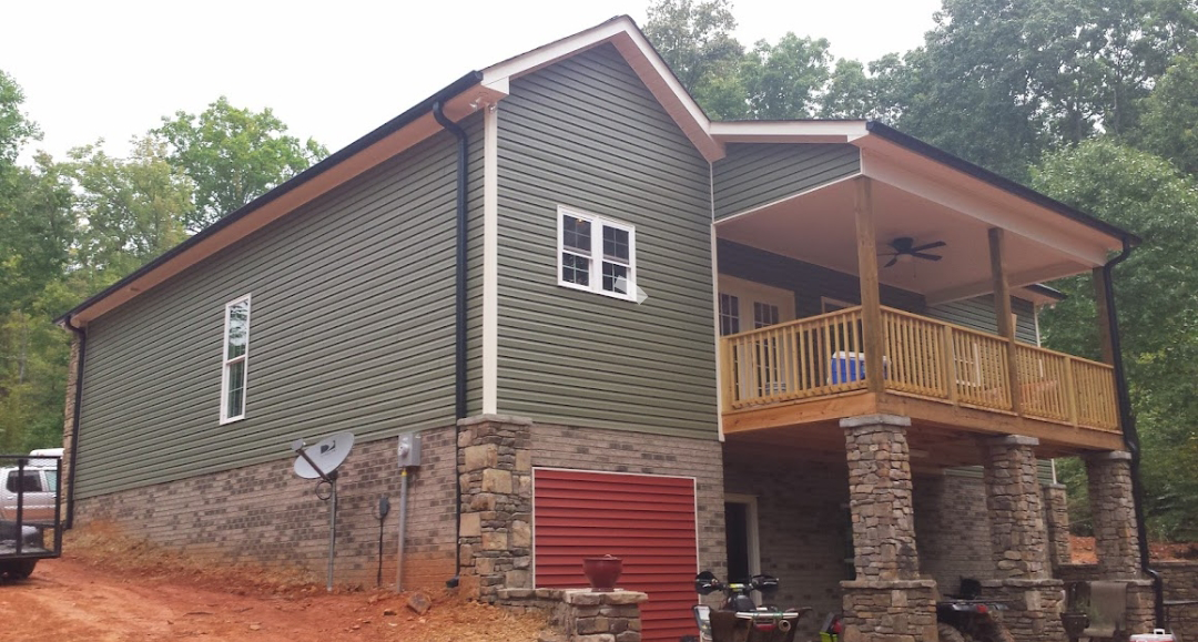Two-story house with green siding and stone foundation, front porch, surrounded by trees.