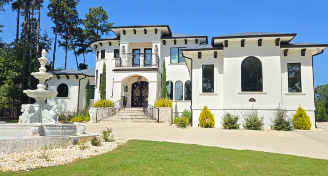 White stucco mansion with arched windows, balcony, and fountain on a sunny day.