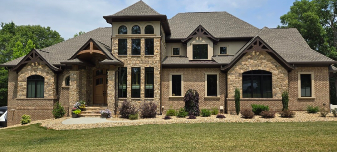Stone-faced, two-story house with a brown roof, set against a green lawn and blue sky.