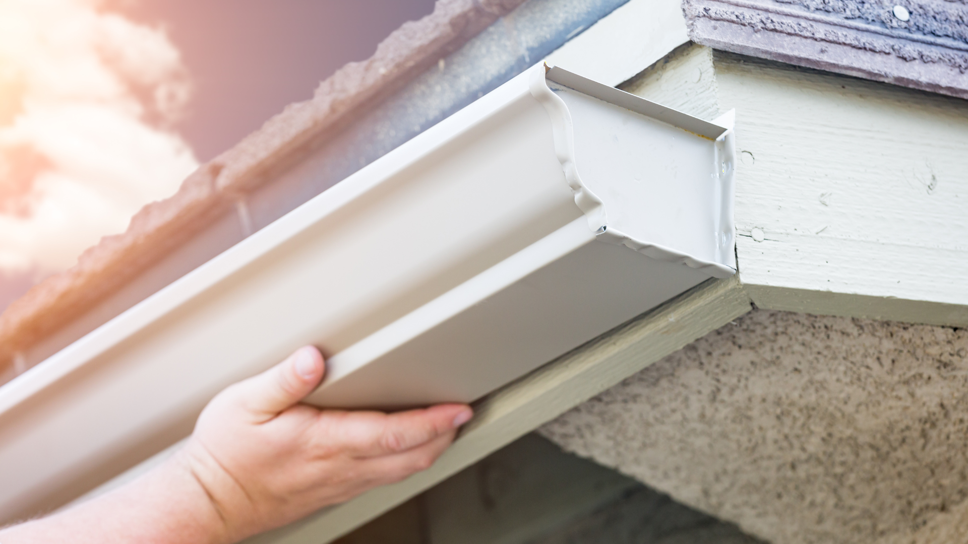 Hand installing a white gutter section on a beige house, near a roof.