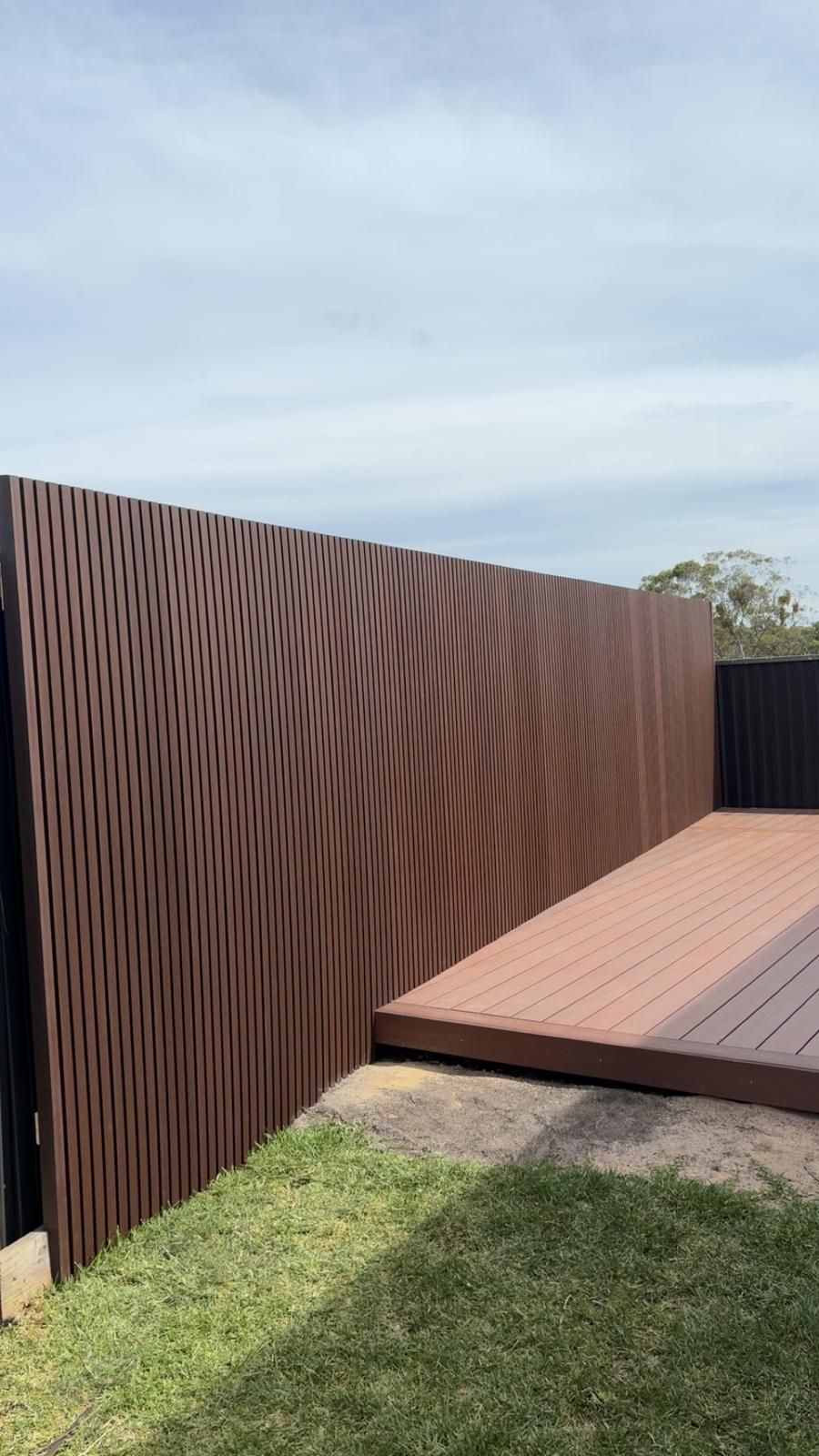 Brown wavy-patterned fence next to a wooden deck and green lawn under a cloudy sky.