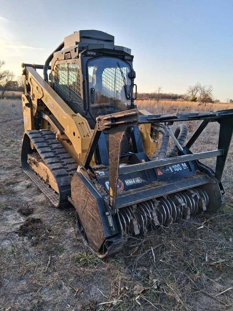 A yellow Caterpillar compact track loader with a forestry mulcher attachment parked in a field during sunset.