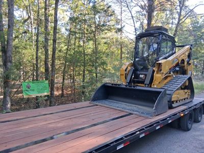 A yellow CAT compact track loader sits on the wooden bed of a flatbed trailer in a forested area.