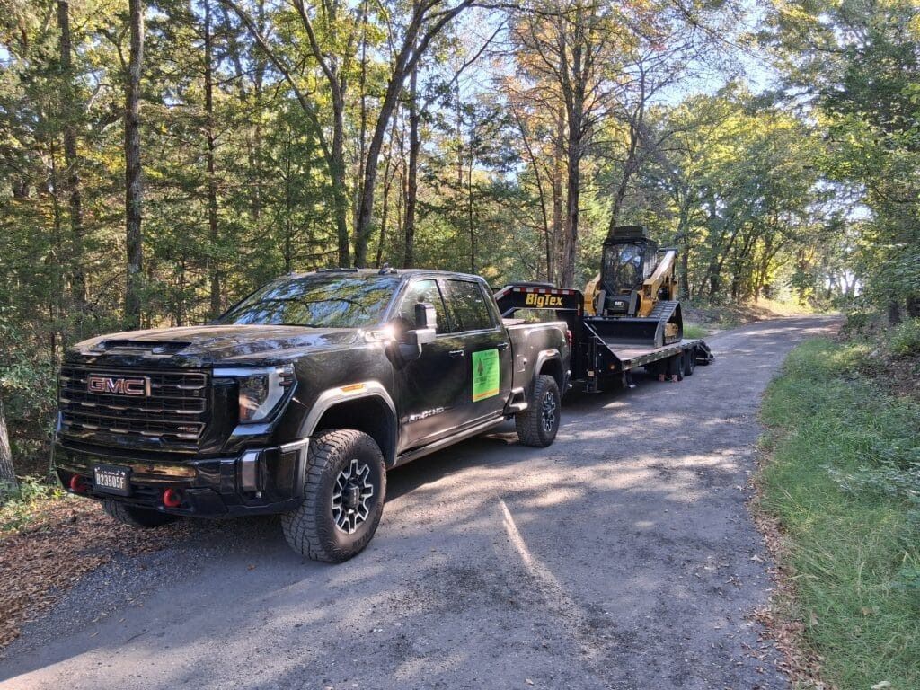 A black GMC pickup truck towing a flatbed trailer with a yellow skid steer on a gravel road through a sunny forest.