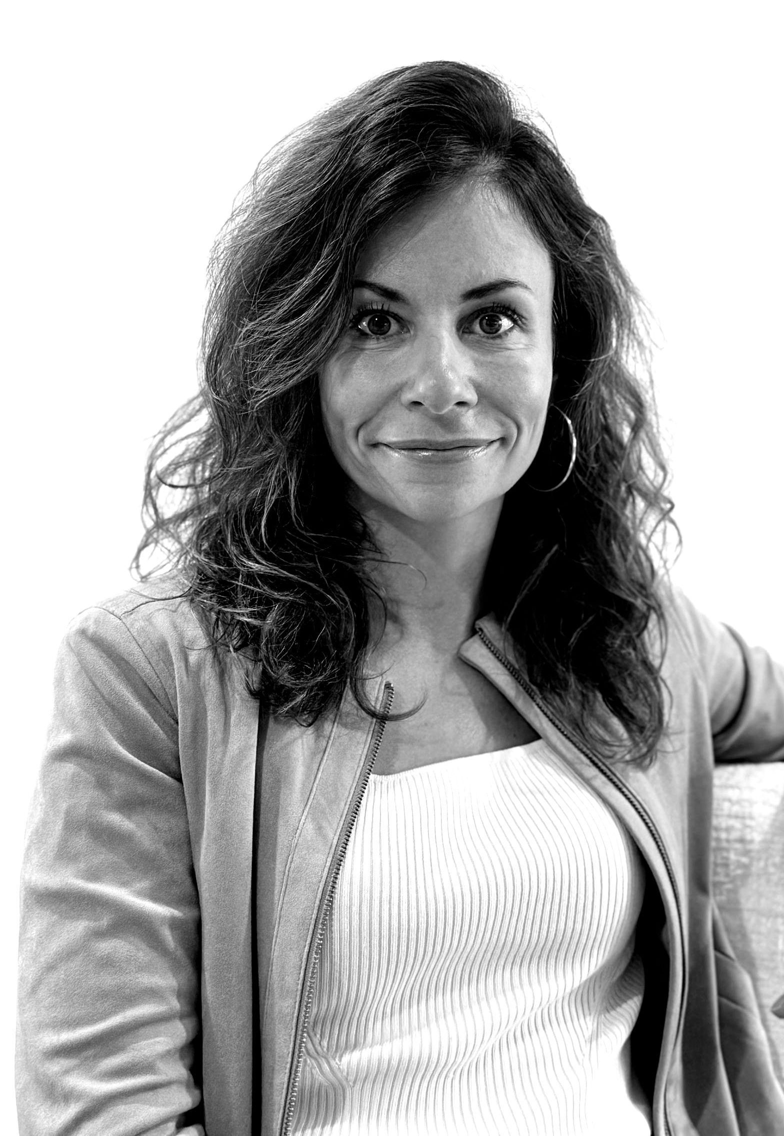Portrait d'une femme souriante aux cheveux bruns bouclés, vêtue d'une veste et d'un haut clairs, sur fond blanc.