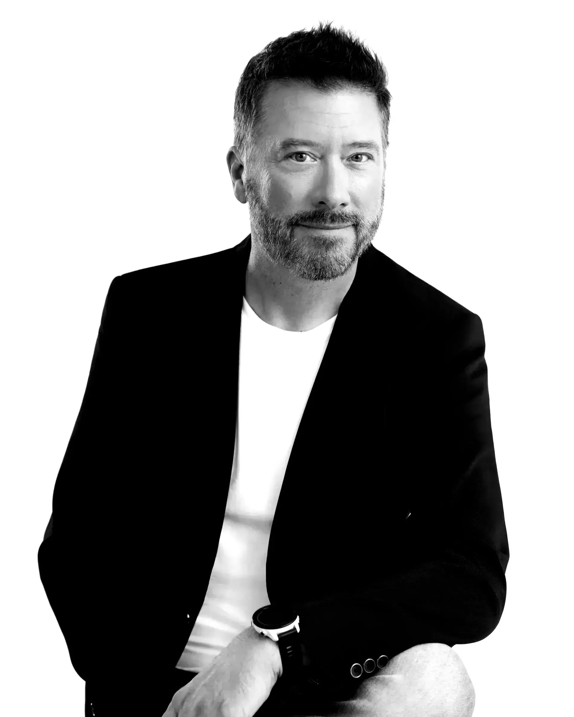 Homme en blazer noir sur chemise blanche, assis et souriant sur fond blanc