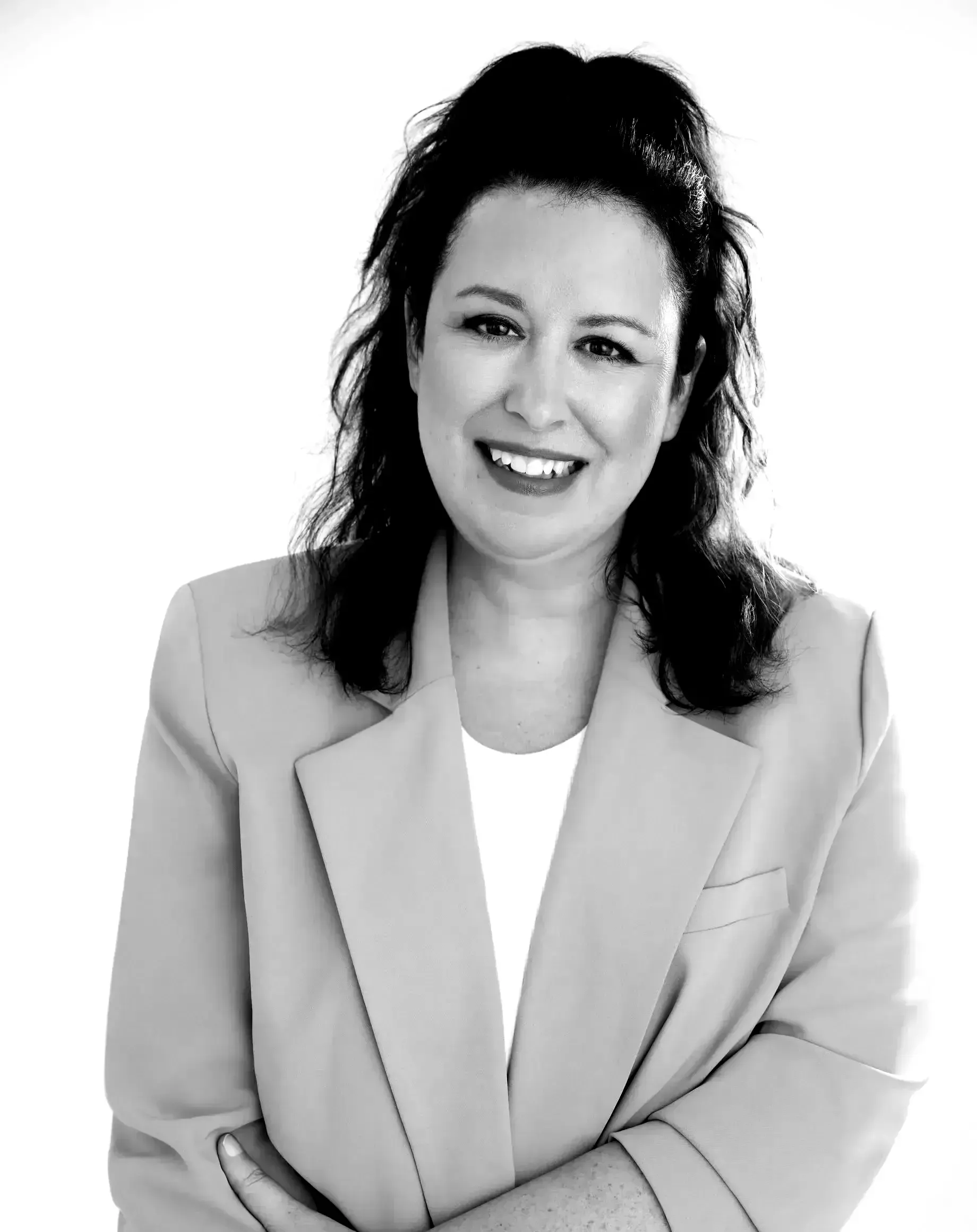 Femme souriante vêtue d'un blazer clair, assise sur un fond uni.