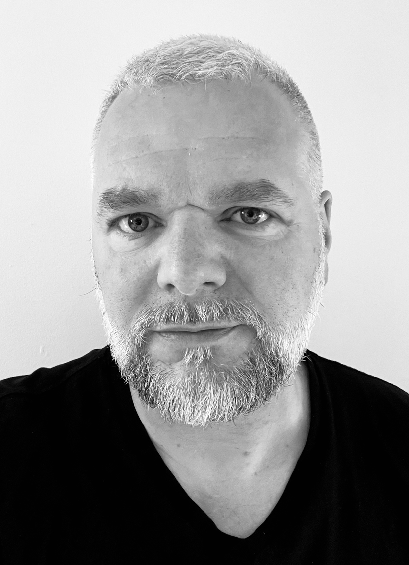 Portrait en gros plan, en noir et blanc, d'un homme aux cheveux gris courts et à la barbe, regardant l'objectif.