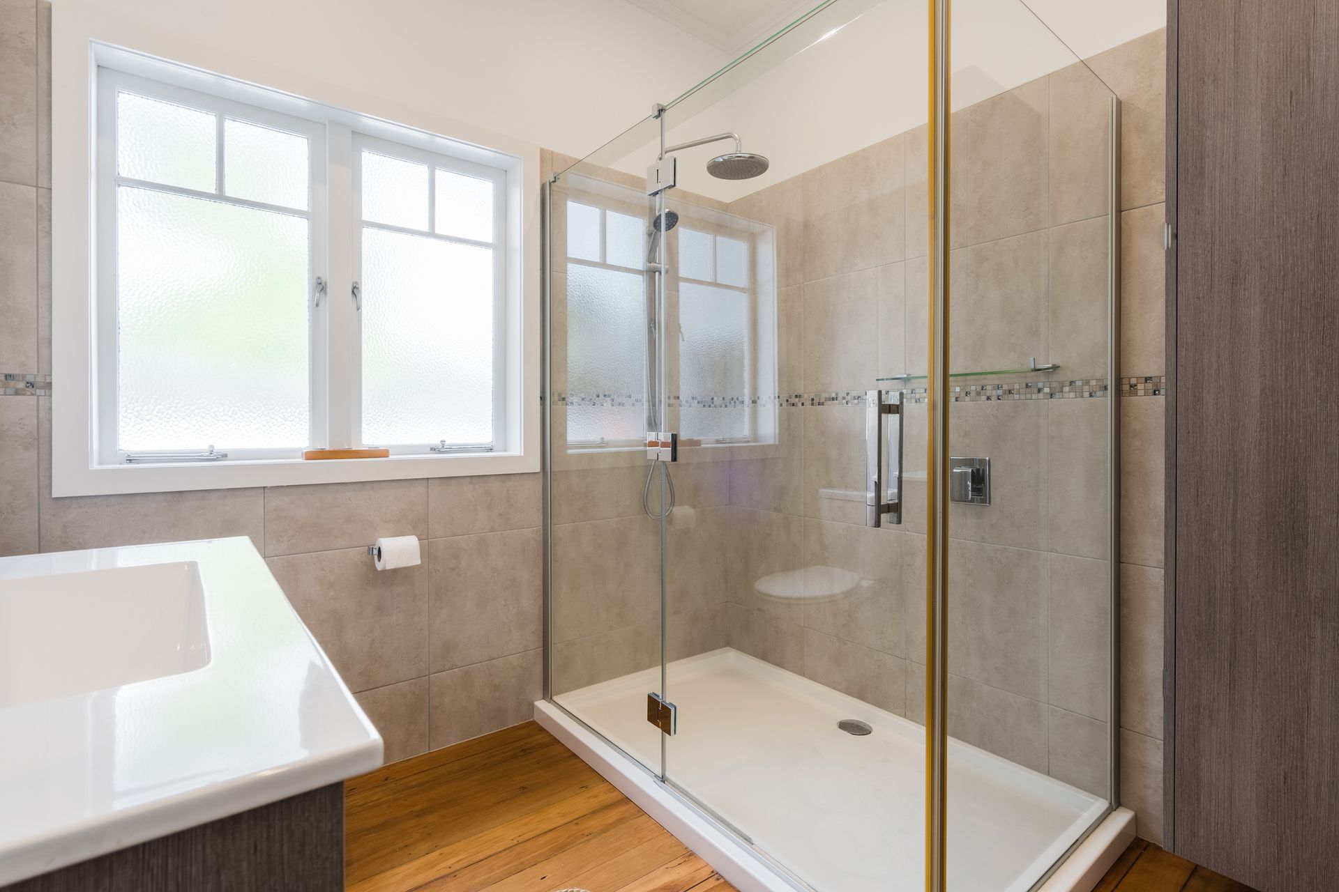 Glass shower with mosaic tiles and rain head in modern bathroom with wood flooring.