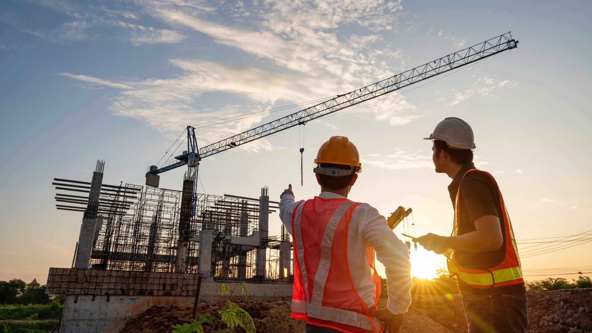 Two construction workers in hard hats at a site with a crane at sunset