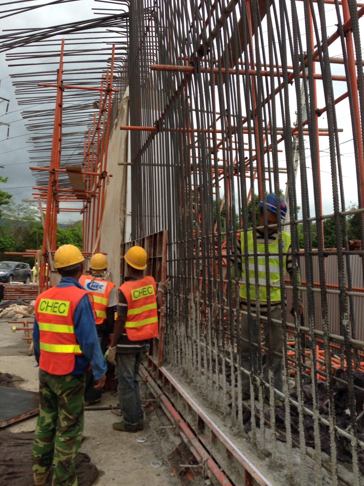 Construction workers in hard hats and orange vests on scaffolding beside a concrete wall reinforcement frame