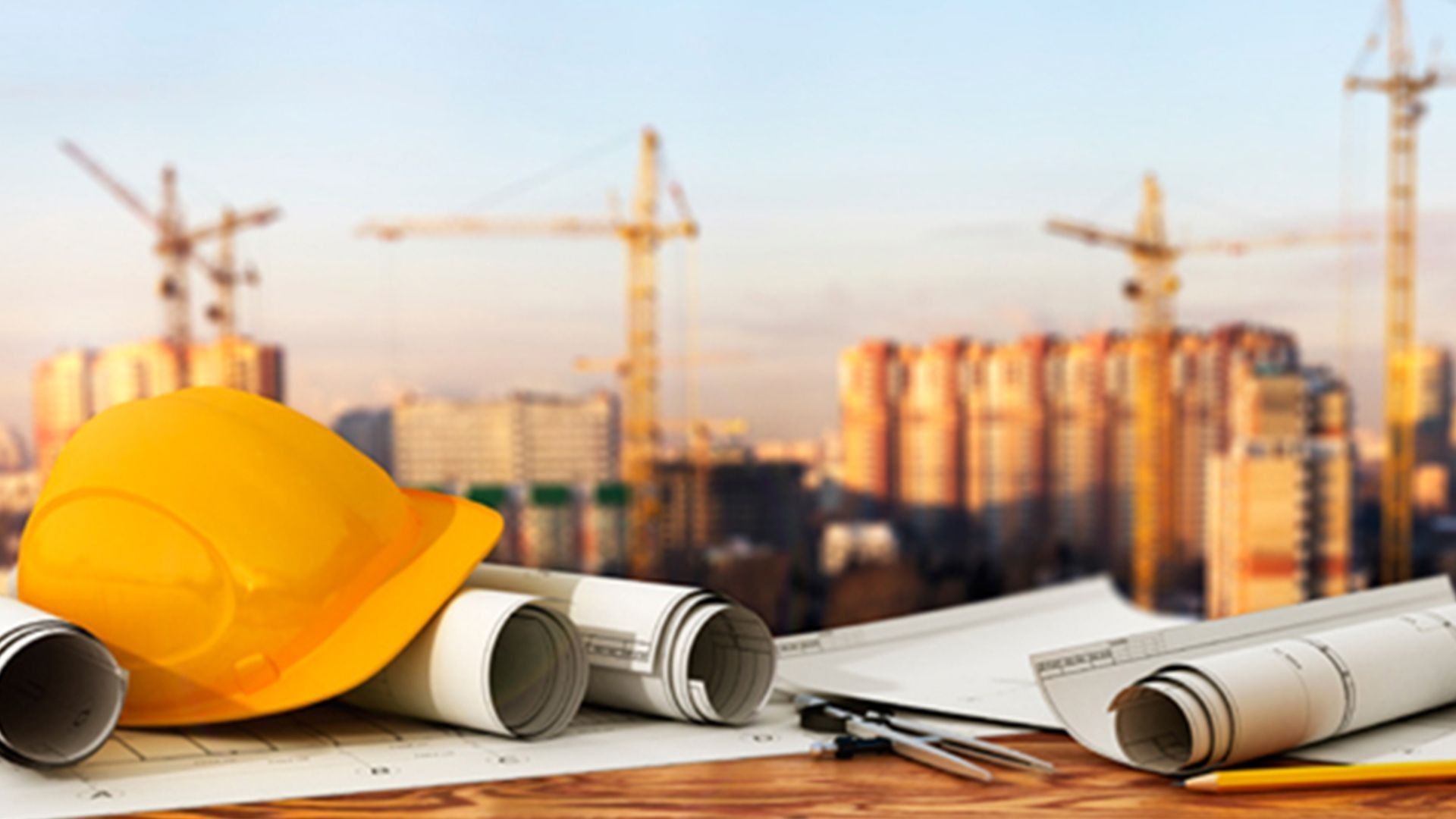 Yellow hard hat and rolled blueprints on a desk, with construction cranes and buildings in the background