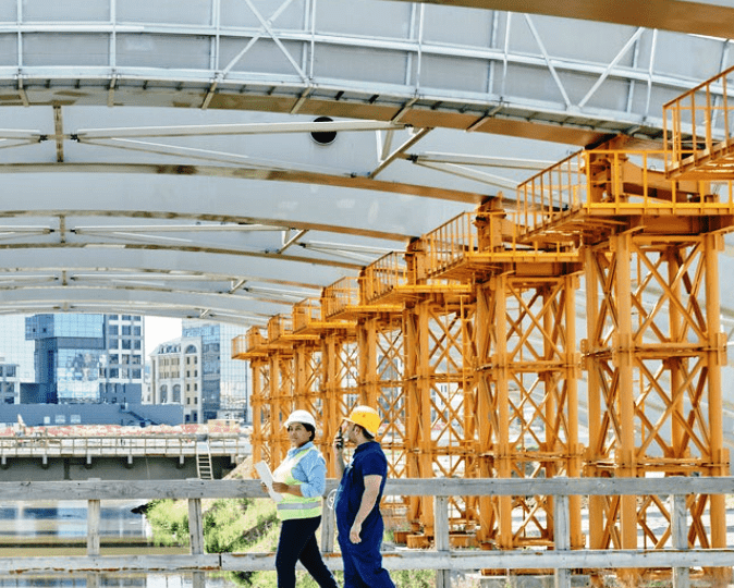 Two workers in hard hats inspecting a bridge under construction with orange scaffolding overhead