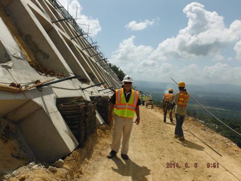 Workers in hard hats and safety vests inspect a damaged hillside building under a cloudy sky.